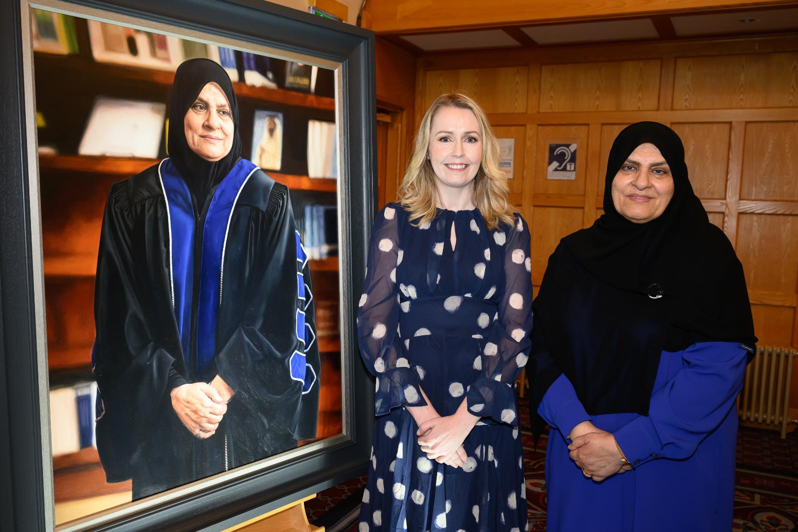 Two women stand posed beside a portrait in a university great hall, looking towards the camera.